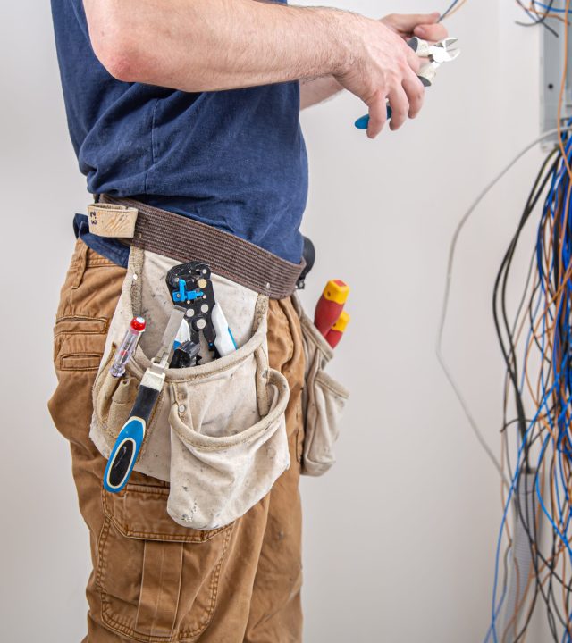 Electrician builder at work, examines the cable connection in the electrical line in the fuselage of an industrial switchboard. Professional in overalls with an electrician's tool.